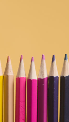 Vertical-Video-Studio-Shot-Of-Multi-Coloured-Pencils-In-A-Line-On-Yellow-Background-